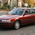 Front three-quarter view of a vintage maroon Mazda 626 midsize sedan parked alongside a curb with autumn leaves