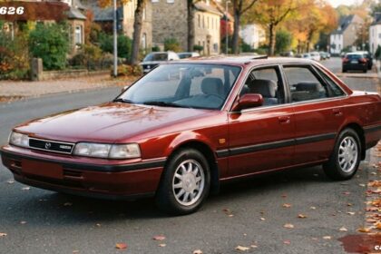 Front three-quarter view of a vintage maroon Mazda 626 midsize sedan parked alongside a curb with autumn leaves