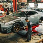 Two mechanics working on a modified grey Nissan GT-R on a hydraulic lift inside an automotive performance shop surrounded by aftermarket car modifications, tools, and brand logos.