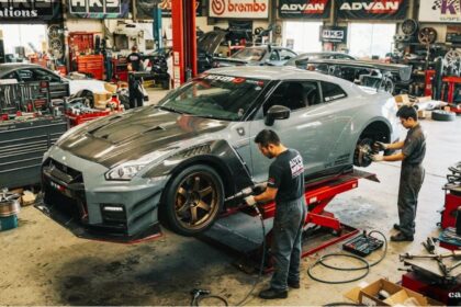 Two mechanics working on a modified grey Nissan GT-R on a hydraulic lift inside an automotive performance shop surrounded by aftermarket car modifications, tools, and brand logos.