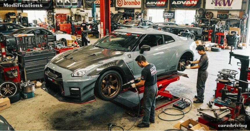 Two mechanics working on a modified grey Nissan GT-R on a hydraulic lift inside an automotive performance shop surrounded by aftermarket car modifications, tools, and brand logos.