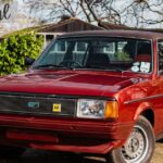 A front three-quarter view of a classic red Morris Ital sedan with a dark vinyl roof parked on a gravel drive with trees and foliage in the background. Cursive text in the top-left corner reads "Morris Ital".