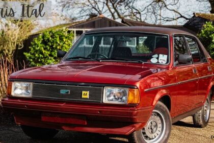 A front three-quarter view of a classic red Morris Ital sedan with a dark vinyl roof parked on a gravel drive with trees and foliage in the background. Cursive text in the top-left corner reads "Morris Ital".