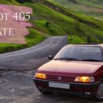 A dark red Peugeot 405 parked on the side of a paved mountain road surrounded by lush green hills. The car's headlights are illuminated, and a purple brushstroke graphic overlay in the top left corner displays the text "PEUGEOT 405 ESTATE" in white.