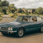 Classic blue-grey Reliant Scimitar GTE parked on a narrow English country road with traditional dry-stone walls and countryside landscape.
