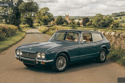 Classic blue-grey Reliant Scimitar GTE parked on a narrow English country road with traditional dry-stone walls and countryside landscape.