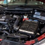 A dirty car air filter exposed inside its open black housing within the engine bay of a red car.