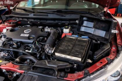 A dirty car air filter exposed inside its open black housing within the engine bay of a red car.