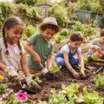 Four young children smiling while planting small green sprouts and watering plants in a raised garden bed outdoors as gardening activites for kids