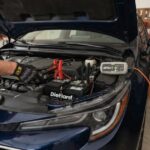A person wearing black mechanic gloves attaches red and black jumper cables from a NOCO Genius 5 battery charger to a DieHard car battery located under the open hood of a blue car.