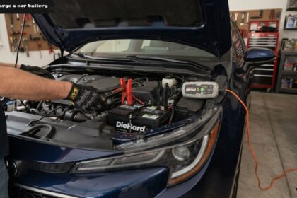 A person wearing black mechanic gloves attaches red and black jumper cables from a NOCO Genius 5 battery charger to a DieHard car battery located under the open hood of a blue car.
