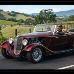 A dark red vintage-style kit car driving along a scenic country road with a couple wearing retro driving goggles and a leather jacket behind the wheel.