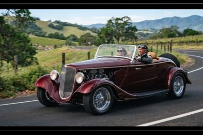 A dark red vintage-style kit car driving along a scenic country road with a couple wearing retro driving goggles and a leather jacket behind the wheel.