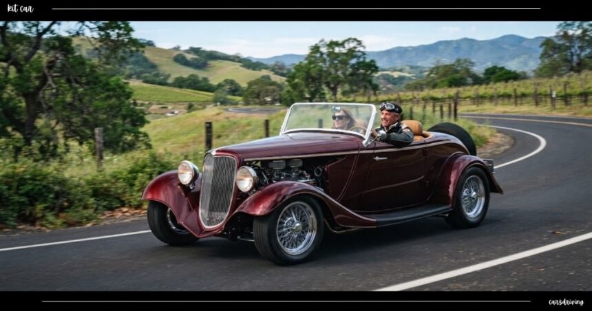 A dark red vintage-style kit car driving along a scenic country road with a couple wearing retro driving goggles and a leather jacket behind the wheel.