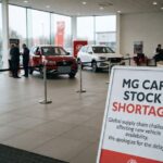 A sign inside a car showroom reading "MG CAR STOCK SHORTAGE" due to global supply chain challenges, with MG vehicles on display in the background.