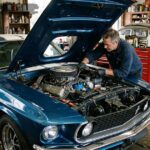 A mechanic working on a classic blue cars with engine exposed under the open hood in a repair shop
