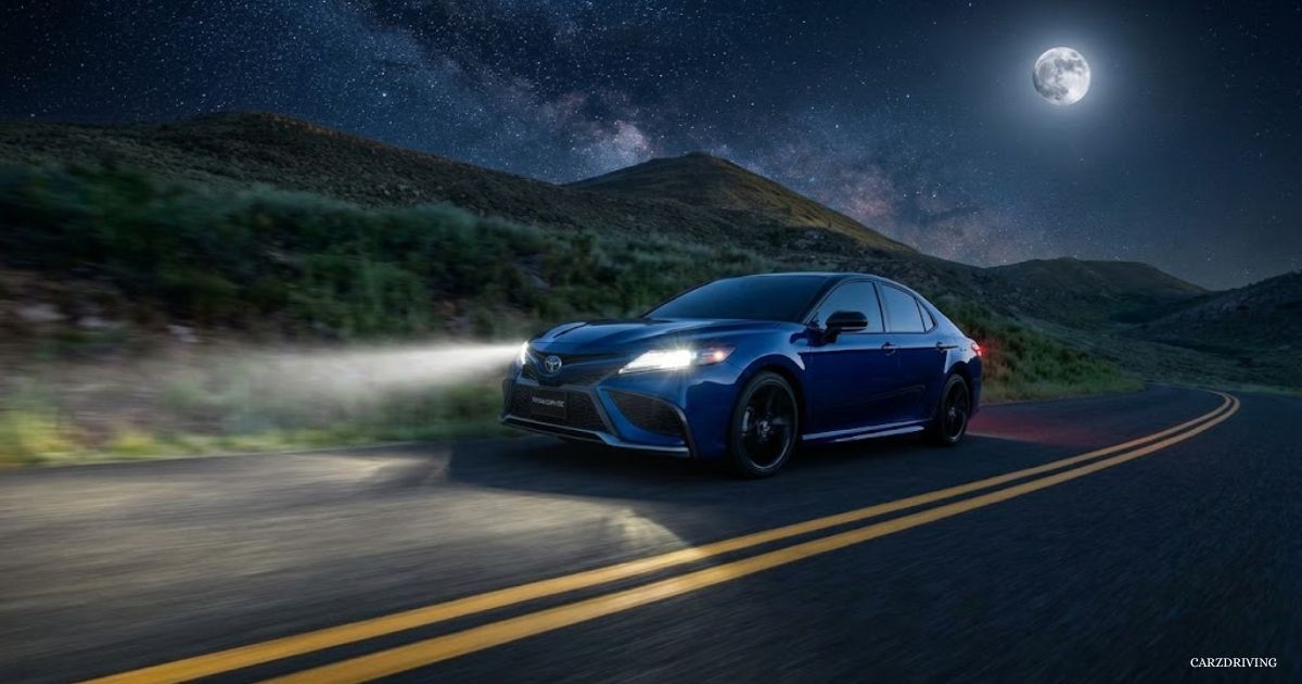 Blue Toyota Camry sedan driving at night on a desert road with headlights illuminated under a starry night sky and full moon.